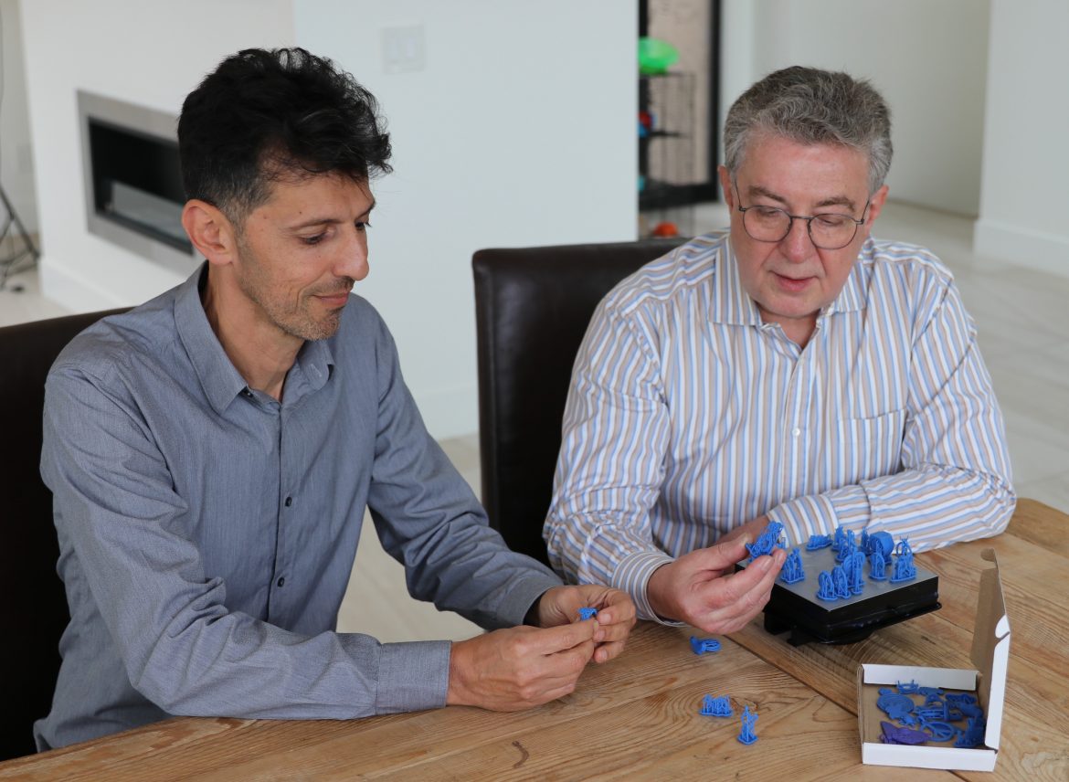 Dror and Aron collaborating over a collection of blue resin 3D-printed prototypes of jewelry pieces on a wooden desk.