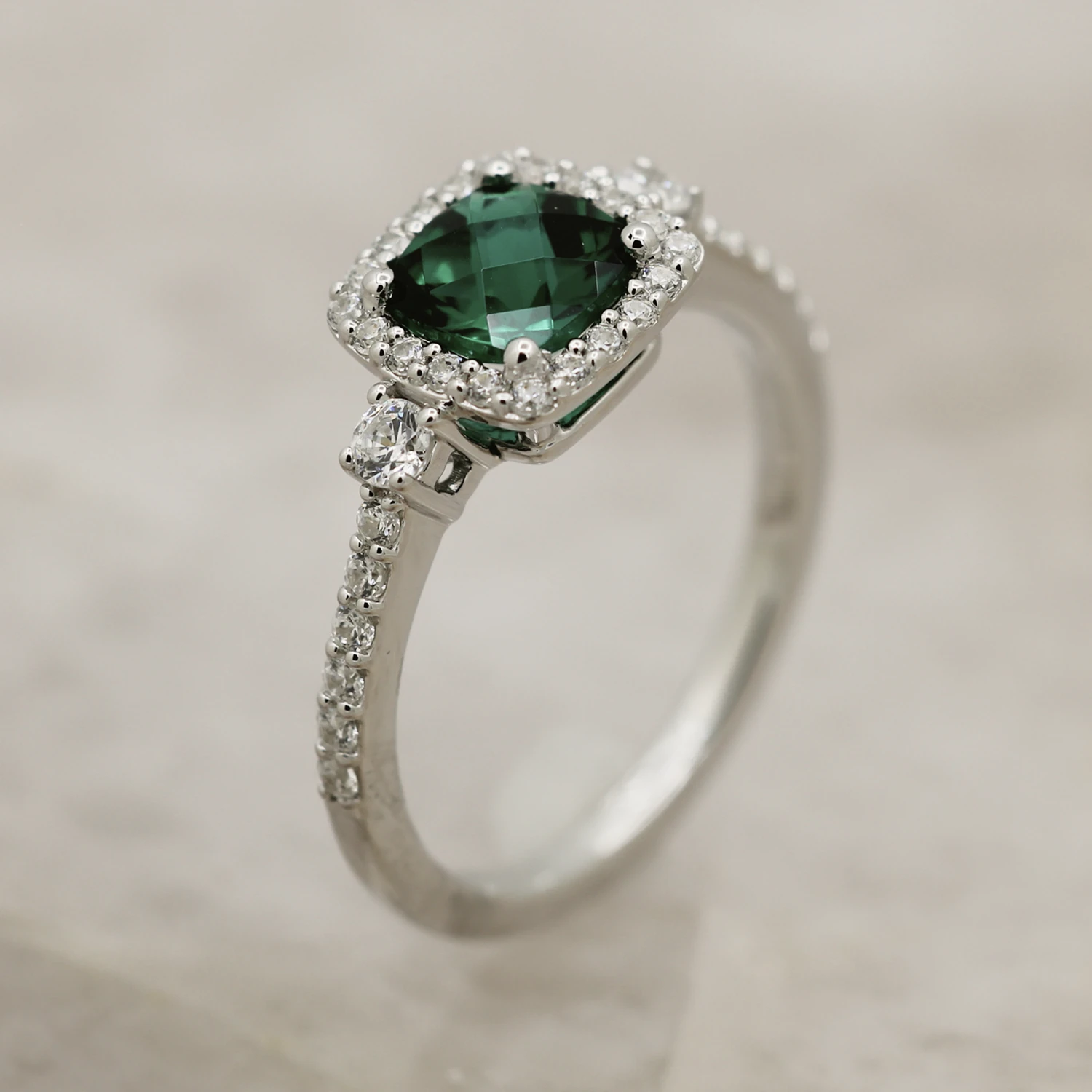 A silver or white gold ring featuring a cushion-cut deep green emerald or tourmaline gemstone surrounded by a diamond halo and a pavé-set diamond band.